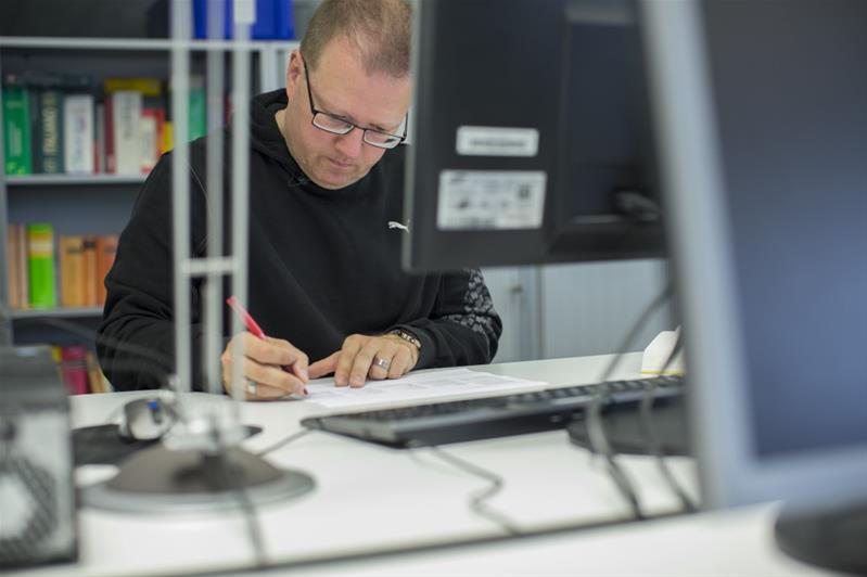 Un uomo davanti a un computer è intento a correggere con una penna rossa un documento stampato.